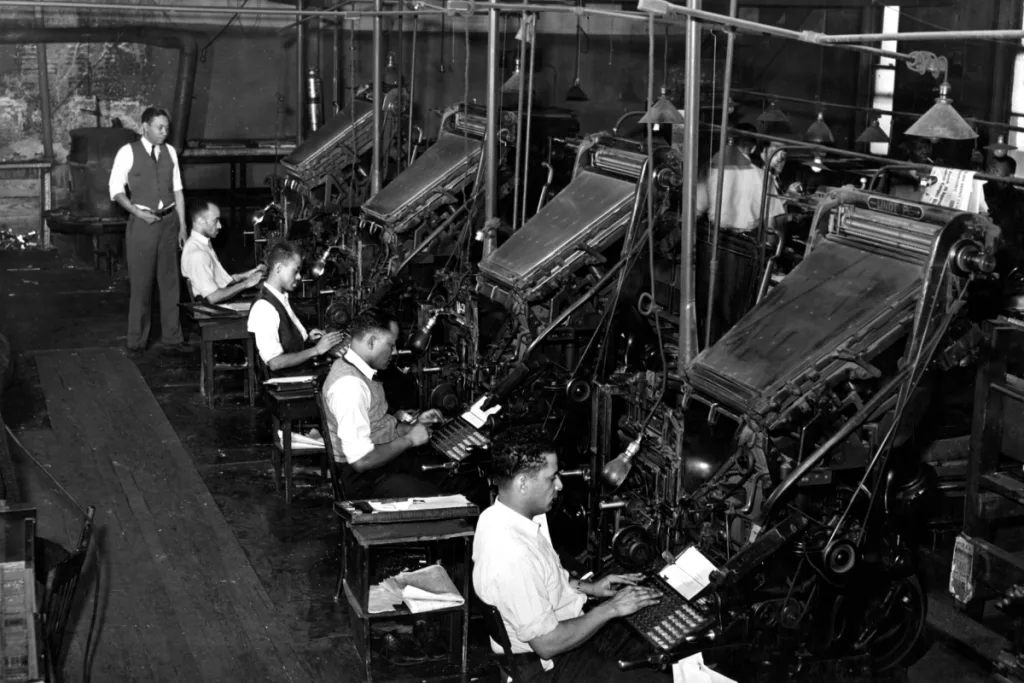 A black and white photo of workers in an assembly line writing on their typewriters, with one man overseeing the operations.
